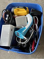 Top view of blue container filled with assorted electric devices including yellow Dorcy flashlight, white grille heater or fan, blue and white handheld iron, headphones, power cords