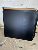 Stack of large black square hardboard or plywood sheets leaning against a wall, smooth surface, approximately 35x33 inches.