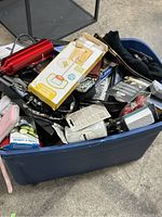 Top-down view showing mixed items inside a large blue plastic bin. Items include a boxed portable mini basketball hoop, stainless steel water bottle, leather wallet, and various packaged electronics and household items.