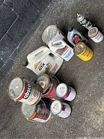 Image of assorted paint cans, motor oil containers, and additives on pavement against a black brick wall.