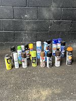 Photo shows 20 aerosol spray cans standing upright against a black brick wall. Various colors and brands visible including yellow, white, green, orange, black, and blue cans.