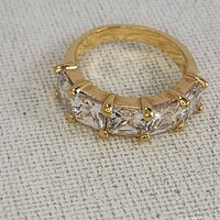 Close-up image of gold-tone ring with five clear square stones, showing detail of prong settings and band texture.