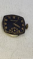 Front view of signed Bulova watch movement with black dial and gold numeral markers, showing winding crown and small seconds dial