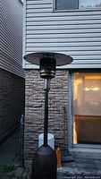 Full view of the outdoor patio propane heater standing next to a brick wall and house exterior, showing height and structure