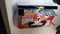 Red and white Pokemon Trading Card Game metal carrying tin with latch, partially open showing edge of booster packs inside.
