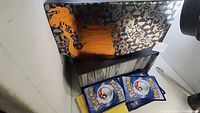 Photo showing a black and orange Pokemon card binder on a white surface with several blue and one yellow Pokemon booster packs placed in front.