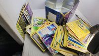 Photo of many loose Pokemon cards stacked and scattered on a surface, with a metal Pokemon card storage tin.