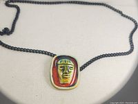 Close-up of the porcelain pendant showing the painted face with green hair, yellow face, and brown-red background on a black chain.