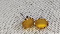 Close-up photo of amber stud earrings showing their oval shape, honey-yellow polished amber stones, and metal scalloped edge settings with post backings.