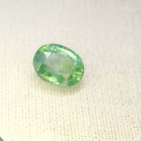 Close-up photo of an oval faceted blue green beryl gemstone on a light fabric background, showing color and cut details.