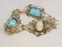 Close-up of antique handmade filigree bracelet with blue topaz stones and cameo centerpiece on a light fabric background.