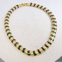 Front view of the necklace showing the alternating black enamel and textured gold-tone segments in a close circle shape on a light fabric background.