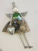Front view of the articulated angel brooch pendant charm showing Swarovski crystal head, metal wings, green fabric bodice with floral sequins, metallic mesh skirt, and heart charm.