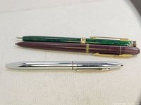 Photo showing three pens: silver-tone, green marbled, and maroon with gold trims placed on a light surface.