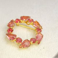 Front angle showing pink teardrop and round rhinestone gems set on gold-tone metal in a circular wreath design on fabric background.