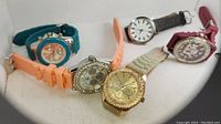 Front view of five fashion watches in multiple colors arranged on white background to show faces and bands.