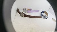 Three watches arranged on white background showing different styles and bands, including brown, lavender, and blue straps.