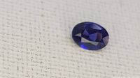 Close-up photo of an oval-cut tanzanite gemstone with deep blue-violet facets on a textured fabric background.
