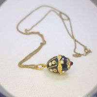 Close-up photo of antique egg pendant on gold-colored chain showing enamel, gem details, and chain clasp.