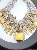 Close-up photo of a large silver-tone chain necklace with gold-tone brutalist geometric accents and a central yellow opalescent square piece.