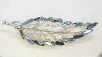 Clear front view of vintage leaf brooch showing silver-tone metal leaf design with blue marquise cut stones lining one edge.