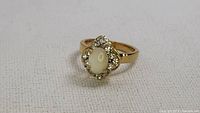 Front view of the vintage gold-tone ring showing the opalescent center stone surrounded by clear rhinestones on a textured white background.