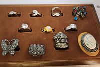Photo showing 11 assorted costume rings on brown display tray. Rings feature rhinestones, enamel, faux pearl, and cameo-style stone.