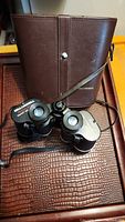 Photo showing Bell and Howell binoculars and original brown leather case lying open on textured surface.