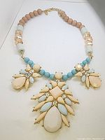 Front view of the large vintage NY signed statement necklace laid flat showing pastel round beads, white and clear faceted beads near the clasp, and three floral pendant drops made of peach and blue stones with rhinestone accents in gold-tone metal.