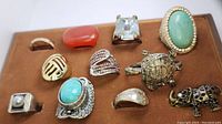 Photo showing 11 different rings on a brown velvet-like tray, including thematic animal shapes and various gemstones and metals.