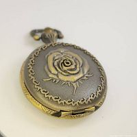 Front view of vintage mini pocket watch showing raised rose motif and ornate border on cover.