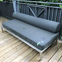 Front view of the outdoor bench showing dark upholstered seat and bolster cushions on a stainless metal frame on a wooden deck. Weathering visible on cushions.