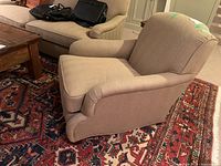 Beige upholstered armchair angled side view on Persian style rug, showing cushioned seat, curved arms, and fabric upholstery.