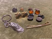 Set of mixed jewelry items on olive cloth background including cufflinks, keychain with sneaker charm, money clip, and pin.
