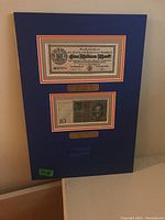 Full view of the framed wall art showing two German banknotes mounted on blue matte board with orange and gray borders and engraved plaque beneath.