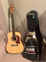 Full view of guitar standing next to soft case, both leaning against wall on carpet