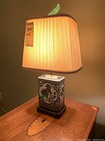 Full view of lamp on wooden side table, ceramic base with blue dragon design and pleated beige cloth shade lit.