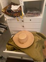 Wide-brimmed woven straw hat on green fabric inside a drawer with folded linens on cabinet above.