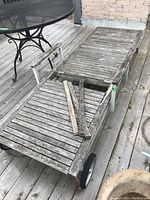 Outdoor wood lounger on metal frame with wheels shown on terrace deck. Visible weathering and broken/missing wooden slats lying on the lounger.