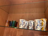 Five decorated Iittala mugs with bird and floral prints, along with five colored drinking glasses in amber, green, blue, red, and orange arranged on a shelf.