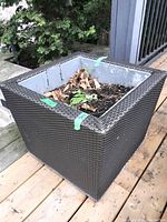 Side view of square woven planter with metal interior showing soil and dried leaves, placed outdoors on wooden deck.