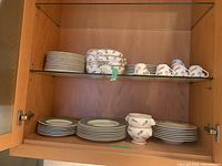Cabinet shelf with stacks of green border and vegetable pattern plates, several vegetable pattern pasta bowls and coffee cups with saucers