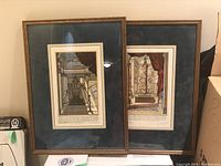 Two framed prints leaning against wall, showing scientific instruments in classical interiors with blue matting and wooden frames.