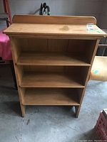 Front view of wooden shelving unit showing three shelves and top flat ledge. Visible stains and scratches on surface. Seen in garage setting.