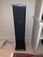 Black floorstanding tower speaker with black cloth grill and plastic base, woodgrain side finish.