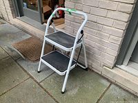 Front angled view of white metal two step folding stool with black rubber steps on outdoor stone patio near brick wall and door.