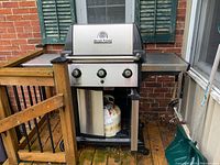 Full view of Broil King Signet gas BBQ grill with propane tank underneath, located outdoors on wooden deck near brick wall.