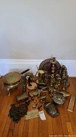 Photo shows an assortment of various brass and cast iron household and decorative items arranged on a wooden floor against a white wall. Includes trays, teapots, candleholders, figurines, and a mirror.