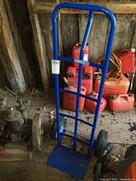 Blue metal hand truck with black wheels and flat base leaning against a wall in a shed.