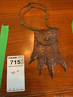 Front view of a textured brown leather coin purse shaped like an alligator or crocodile foot with black claw tips, snap fastener, and attached cord strap on a wooden surface.